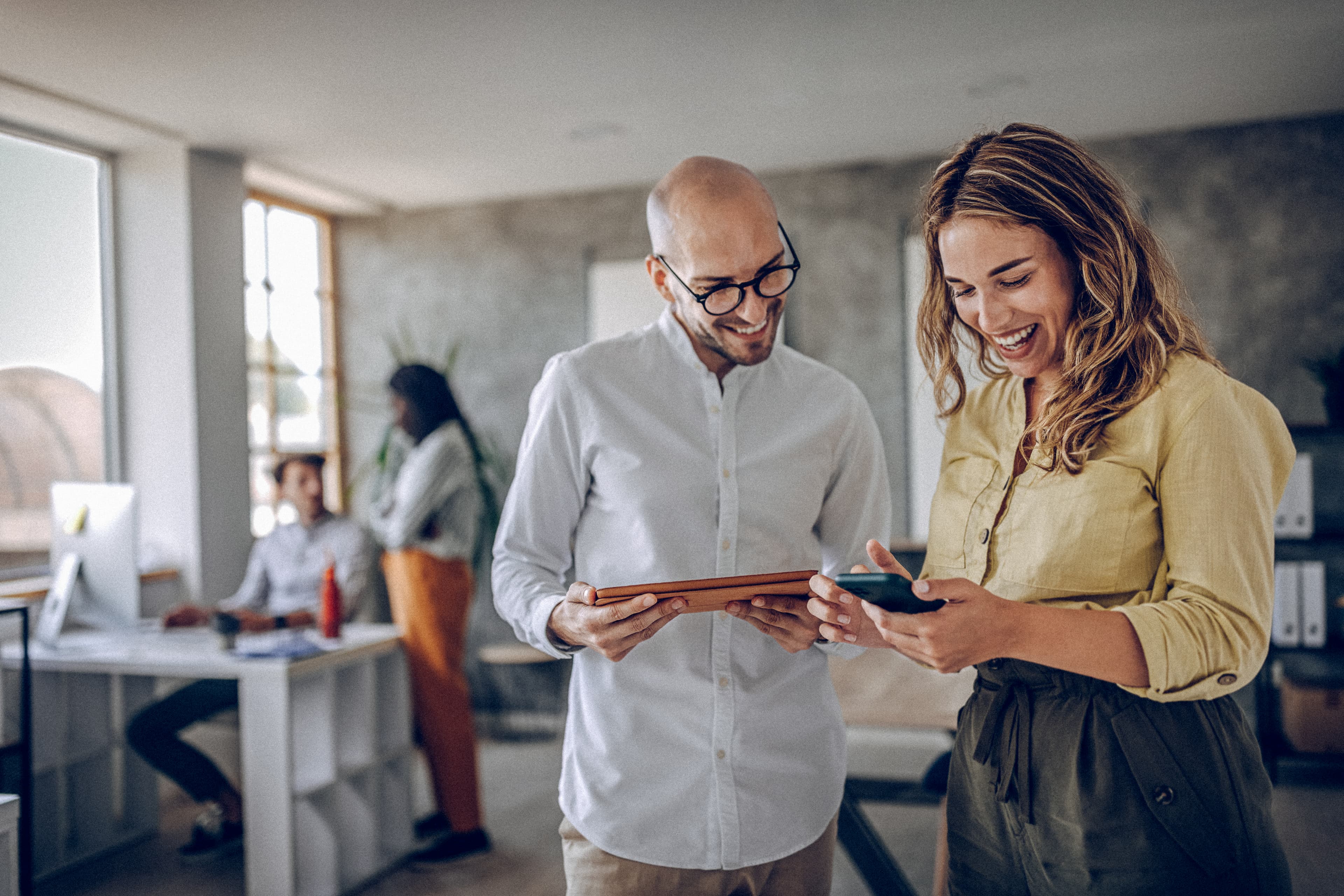 A man and woman in an office, smiling as they engage with an iPad and phone, highlighting a collaborative and positive work environment focused on digital connectivity.