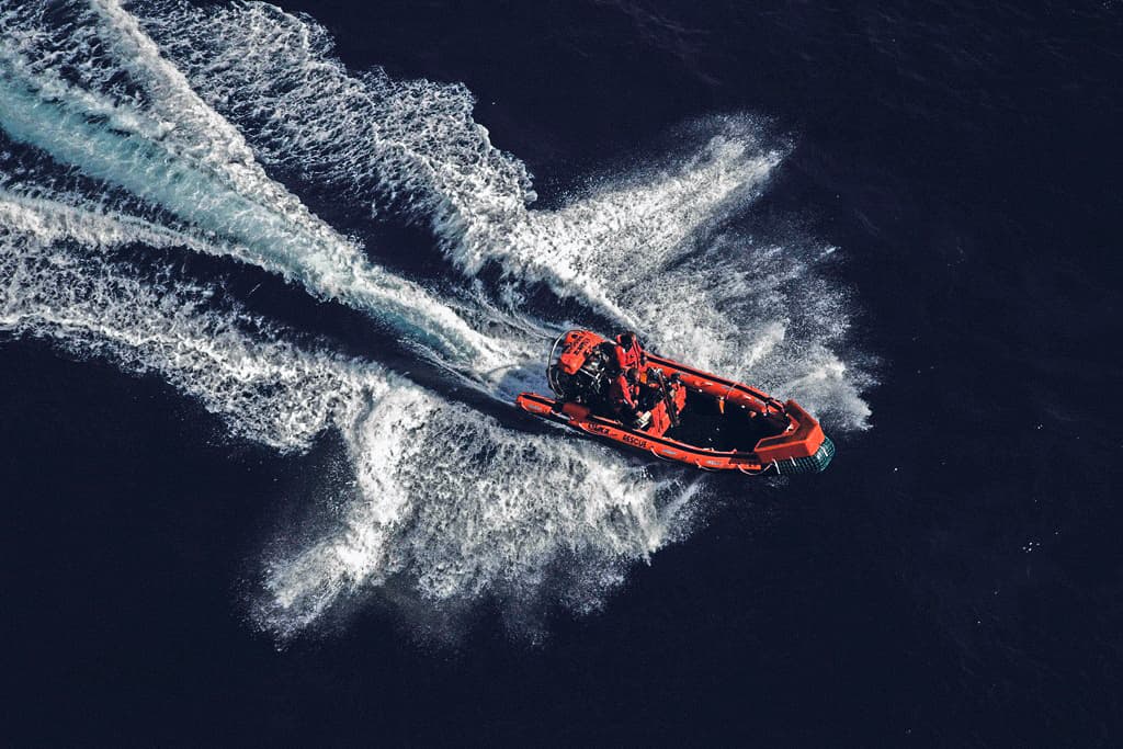 The image shows an orange rescue boat navigating through dark blue waters, leaving white, foamy trails behind. The boat’s speed creates dynamic water patterns, emphasising swift movement and control.