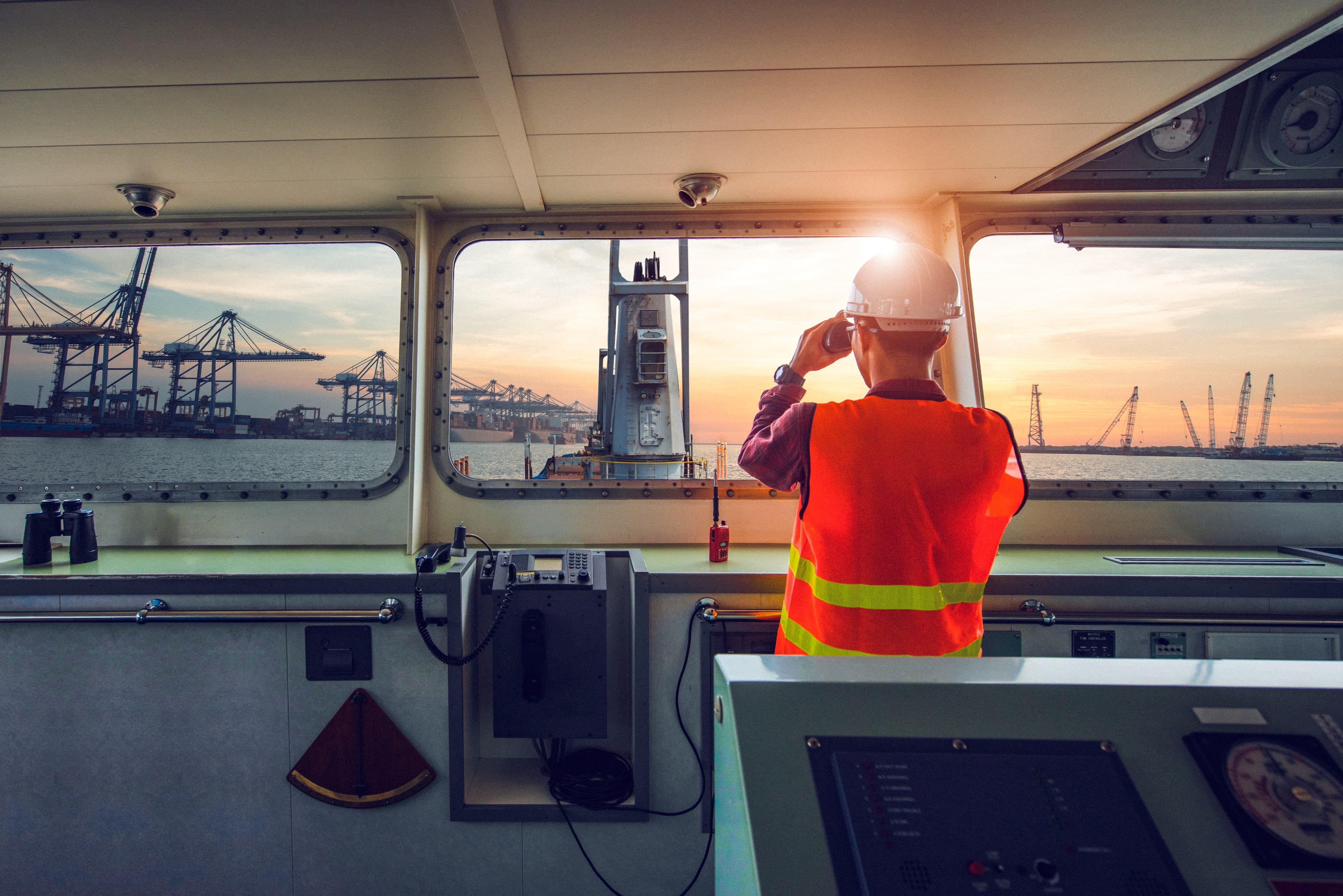 A maritime worker wearing a high-visibility vest looks out through the bridge of a vessel, observing port cranes in the distance, illustrating coordination and oversight in port operations.