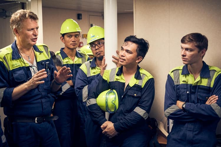 A group of workers in protective uniforms and helmets gather around a senior team member, who is explaining or instructing something. The crew listens intently, reflecting teamwork and knowledge sharing in a maritime setting.