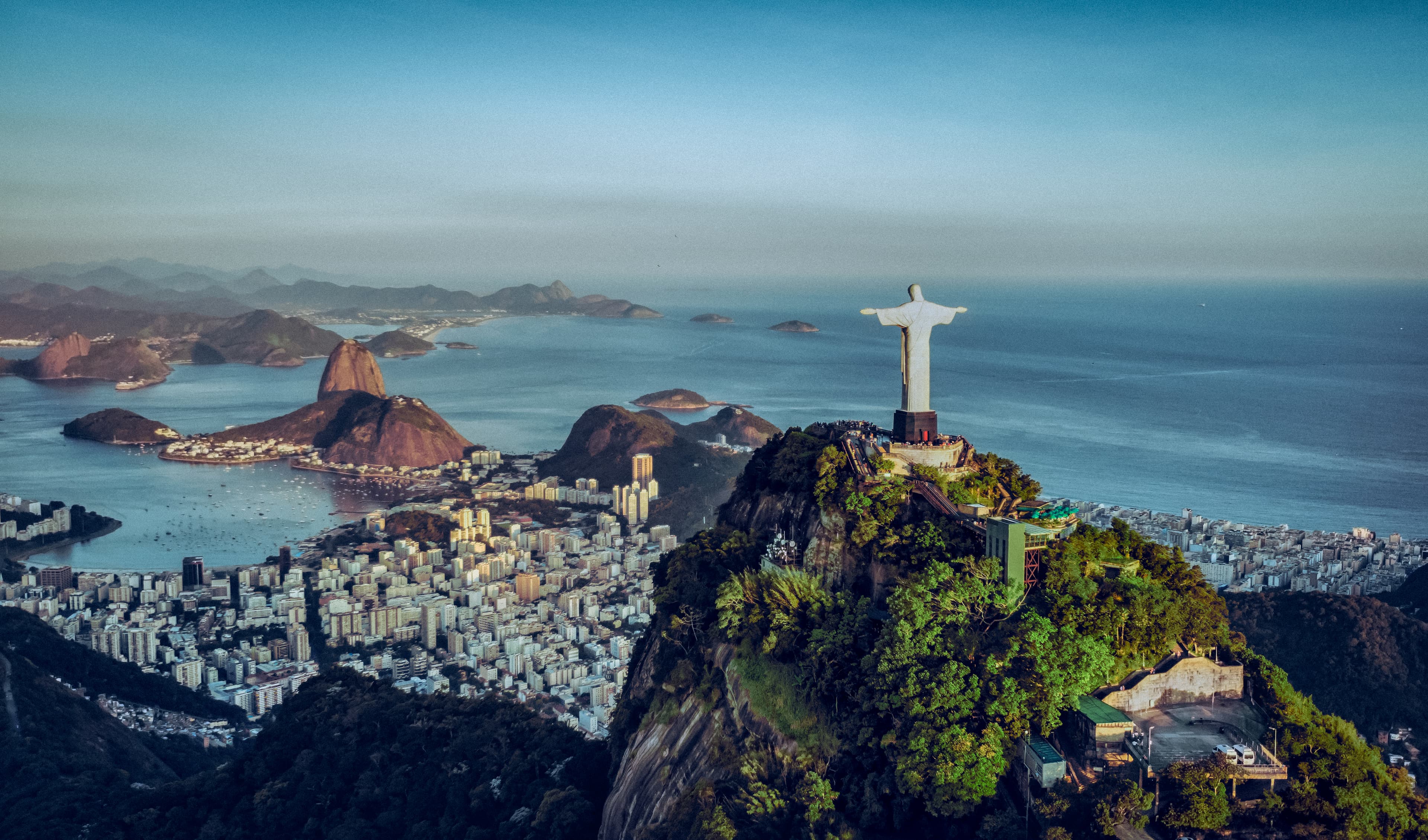 A stunning aerial view of Rio de Janeiro, featuring the iconic Christ the Redeemer statue overlooking the city’s coastline, mountains, and Sugarloaf Mountain, showcasing Brazil’s breathtaking natural beauty and landmarks.