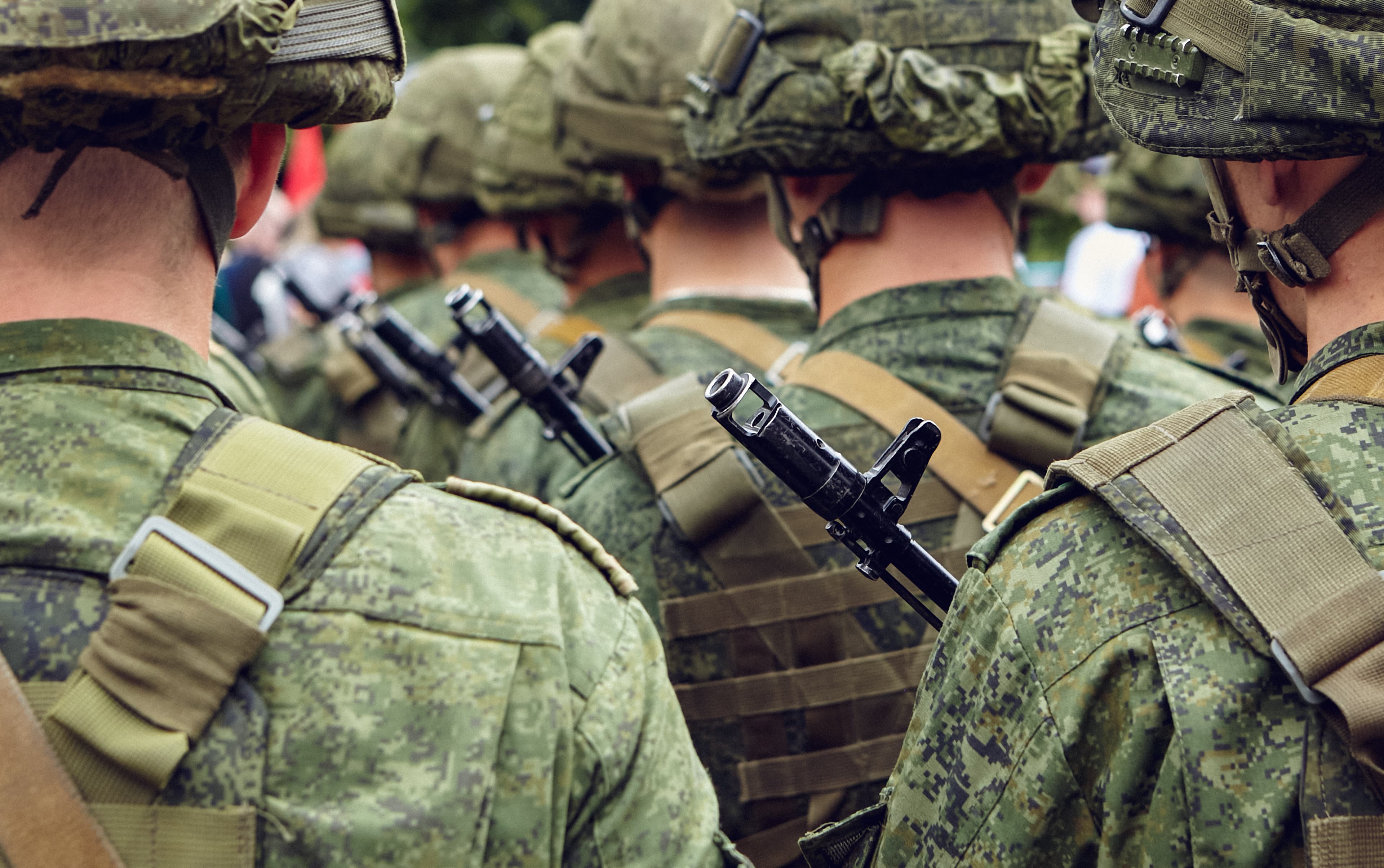 Group of soldiers in camouflage uniforms marching in formation, viewed from behind with rifles strapped to their backs.